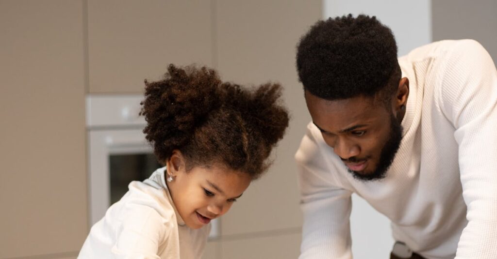Glad young bearded African American father spending time with cute little daughter sitting on table with bowl of tasty ripe fruits in kitchen