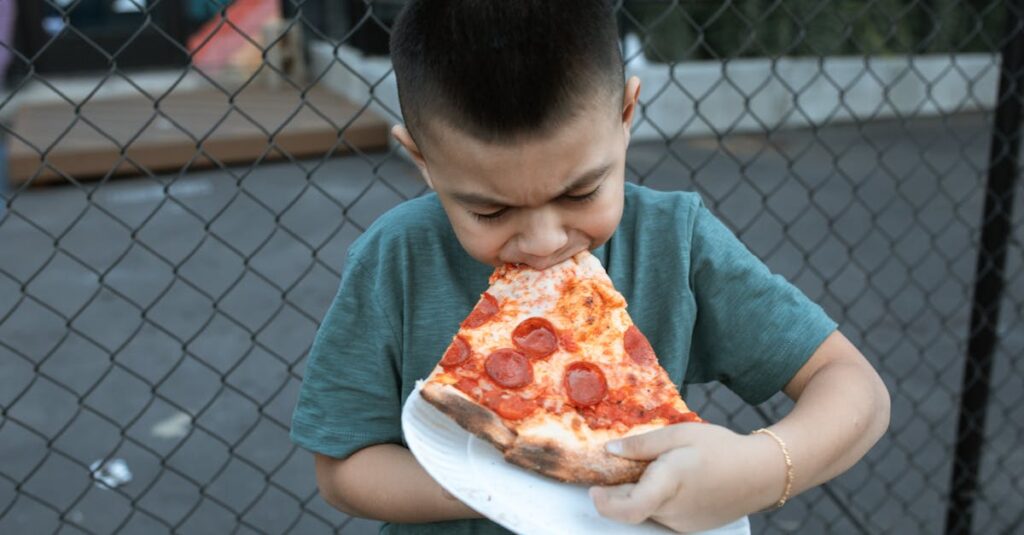 A child in a green shirt eagerly bites into a large pepperoni pizza slice outdoors.